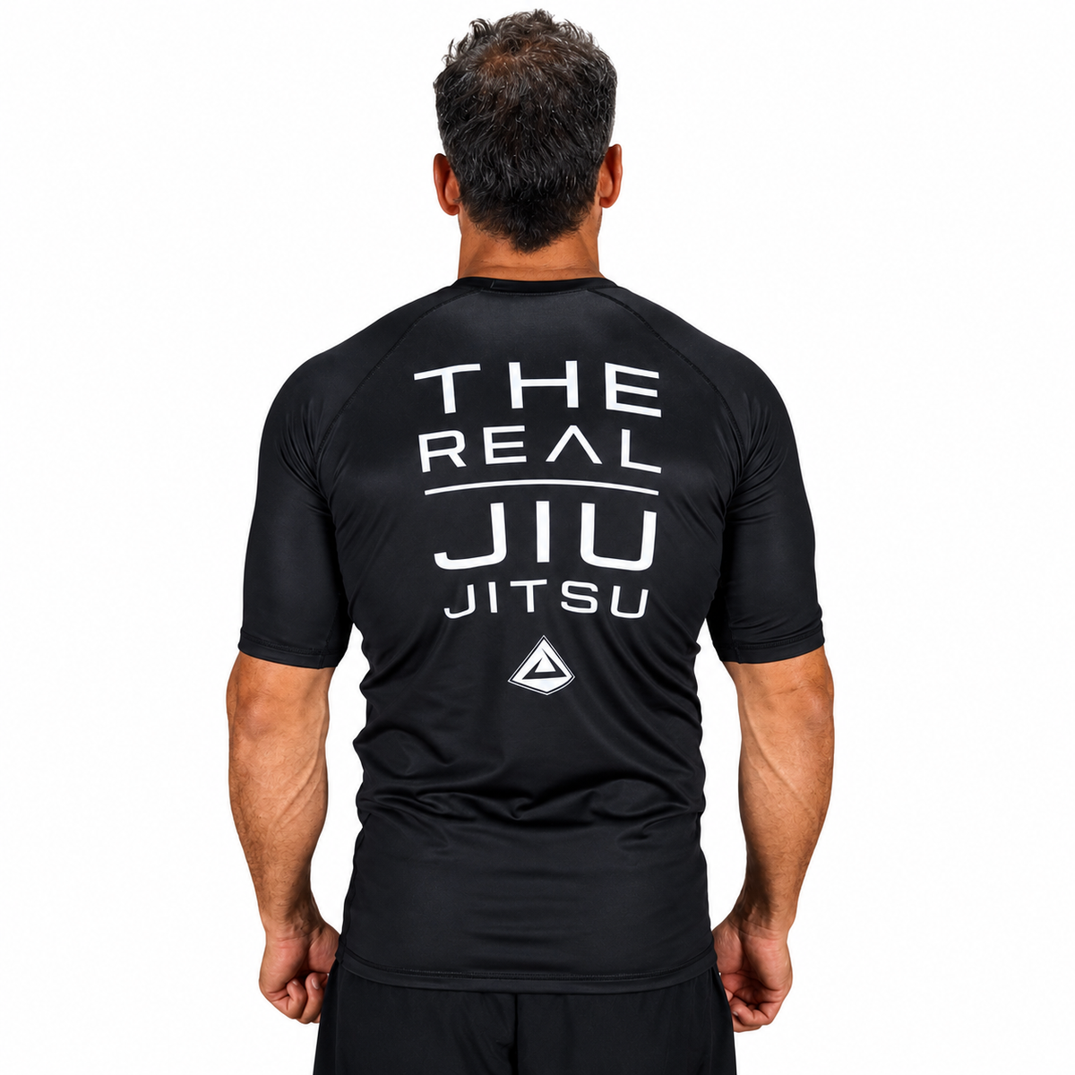 A man viewed from behind wearing a black short-sleeve athletic shirt with large white text on the back reading 'THE REAL JIU JITSU' and a small triangular logo below, against a plain white background.
