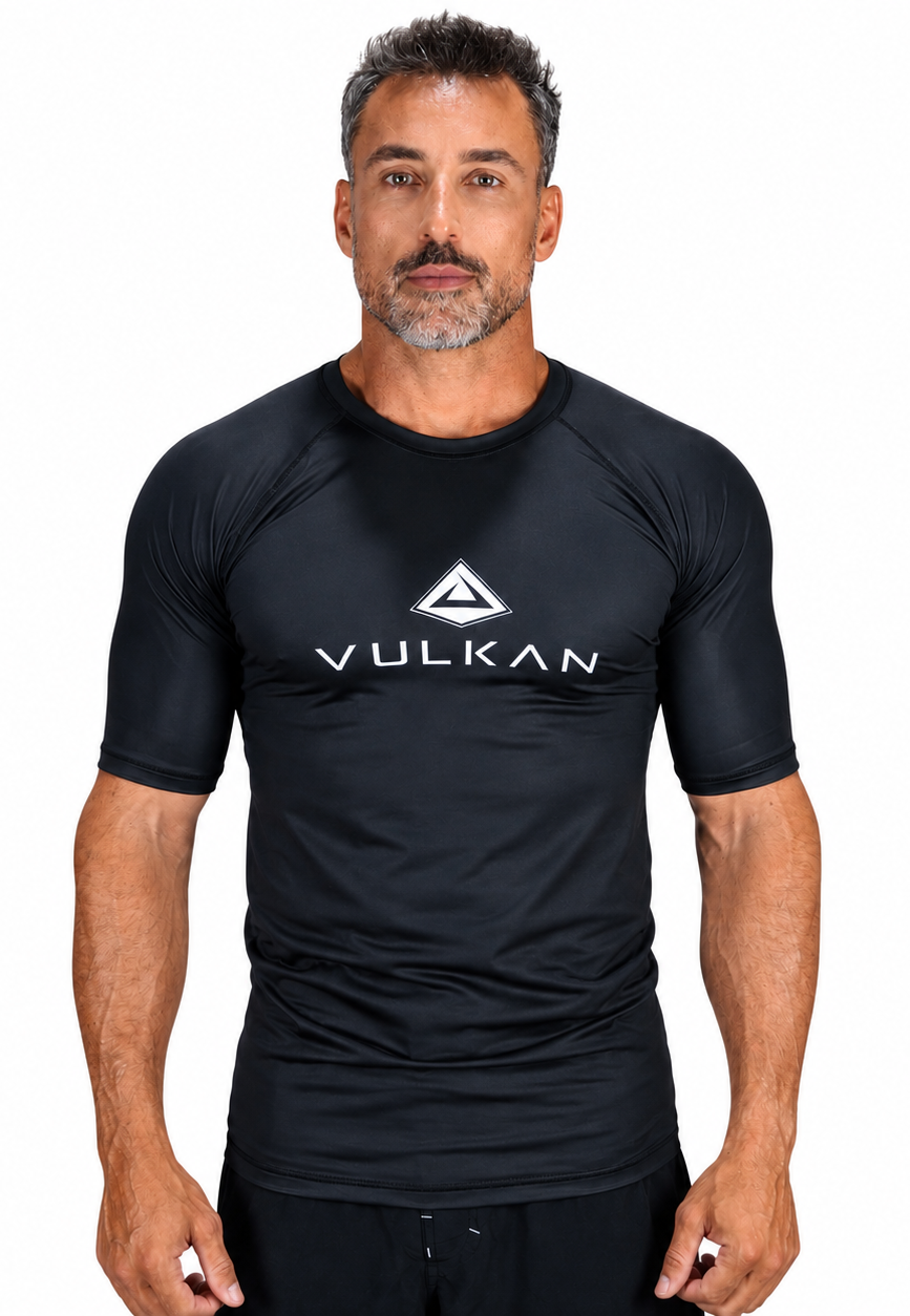 A man wearing a black short-sleeve athletic rash guard with a white VULKAN logo printed on the chest, set against a plain white background.